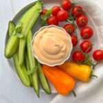 A plate with sliced cucumbers, whole cherry tomatoes on the vine, two yellow mini bell peppers, and a bowl of creamy dipping sauce—perfect for pairing while you learn how to make spicy mayo—arranged neatly on a white surface with a white napkin.