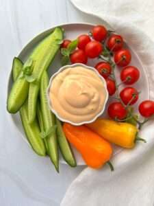 A plate with sliced cucumbers, whole cherry tomatoes on the vine, two yellow mini bell peppers, and a bowl of creamy dipping sauce—perfect for pairing while you learn how to make spicy mayo—arranged neatly on a white surface with a white napkin.