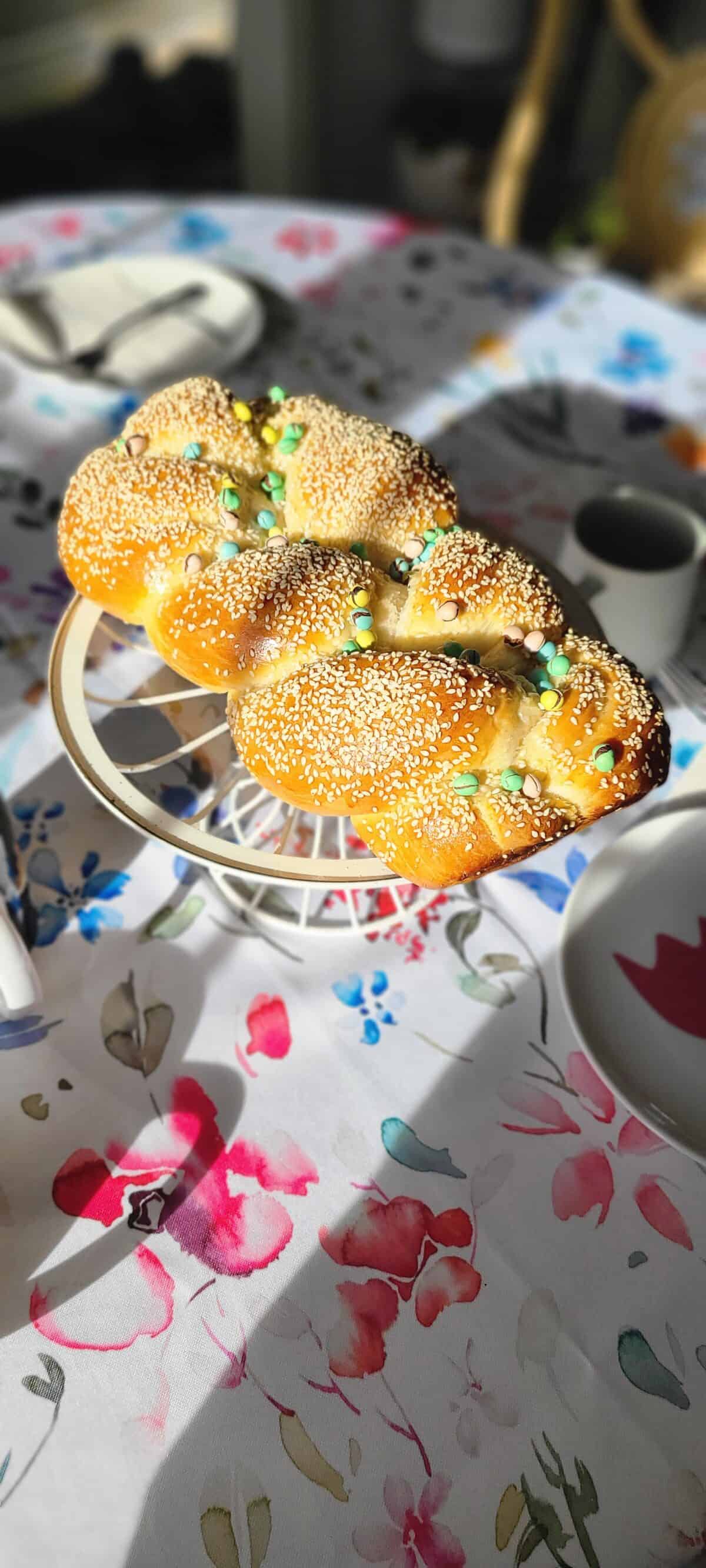 A braided loaf of bread topped with sesame seeds and colorful candy pearls sits on a white stand, placed on a floral-patterned tablecloth in bright sunlight.
