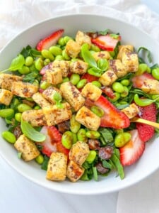 A bowl of The Kourtney Kardashian Salad with cubed tofu, sliced strawberries, edamame, greens, dates, and fresh basil leaves, artfully arranged on a white surface.