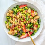 A bowl of Ariana Grande Salad with leafy greens, cubed tofu, sliced strawberries, edamame, and a drizzle of dressing. A gold fork rests inside the bowl, with a white cloth nearby.