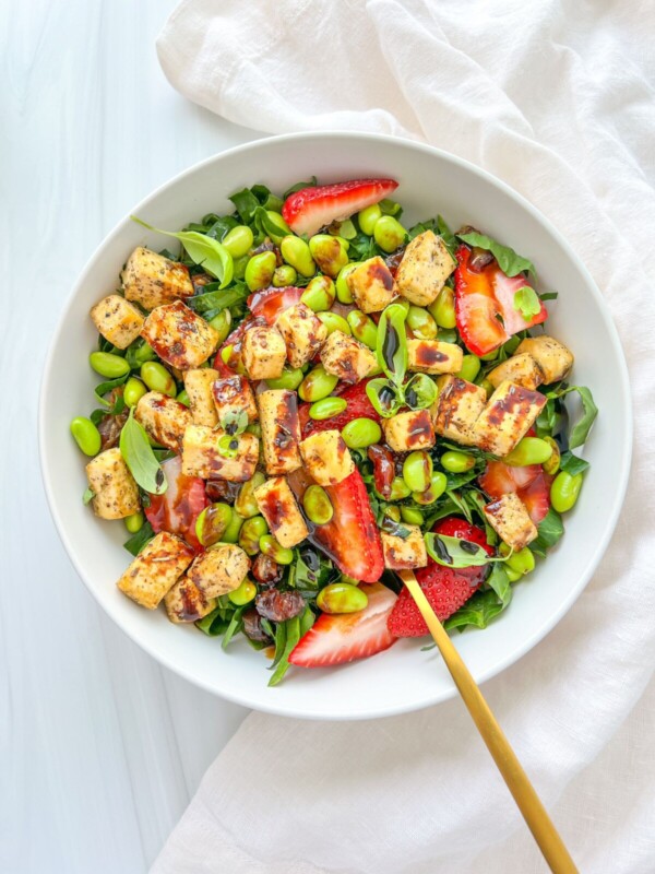 A bowl of Ariana Grande Salad with leafy greens, cubed tofu, sliced strawberries, edamame, and a drizzle of dressing. A gold fork rests inside the bowl, with a white cloth nearby.