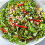 A white bowl filled with Ariana Grande Salad—arugula topped with sliced red peppers, chopped nuts, shaved cheese, and a drizzle of balsamic glaze. A fork rests on the side of the bowl.