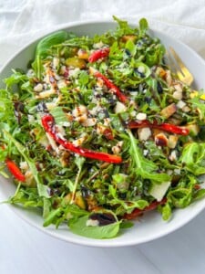 A white bowl filled with Ariana Grande Salad—arugula topped with sliced red peppers, chopped nuts, shaved cheese, and a drizzle of balsamic glaze. A fork rests on the side of the bowl.
