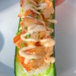 A hand holds a halved cucumber transformed into a Cucumber Sushi Boat, filled with rice, topped with seasoned salmon, creamy sauce, and chopped green herbs, served on a white plate.