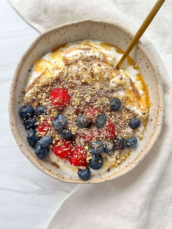 A refreshing Yogurt Bowl topped with strawberries, blueberries, seeds, and honey, with a spoon resting inside. The bowl sits on a light cloth on a white surface.