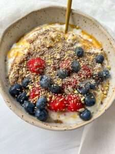 A nourishing Yogurt Bowl topped with fresh blueberries, strawberries, flaxseeds, sunflower seeds, and bee pollen, drizzled with honey and served with a spoon.