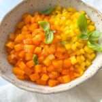 A bowl filled with small, neatly diced orange and yellow bell peppers, garnished with fresh basil leaves, sits on a light-colored cloth—perfect for showcasing your skills in How To Dice Bell Peppers.