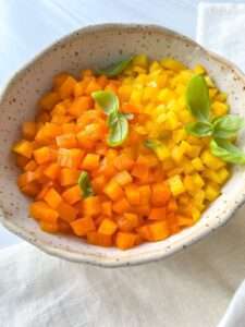 A bowl filled with small, neatly diced orange and yellow bell peppers, garnished with fresh basil leaves, sits on a light-colored cloth—perfect for showcasing your skills in How To Dice Bell Peppers.