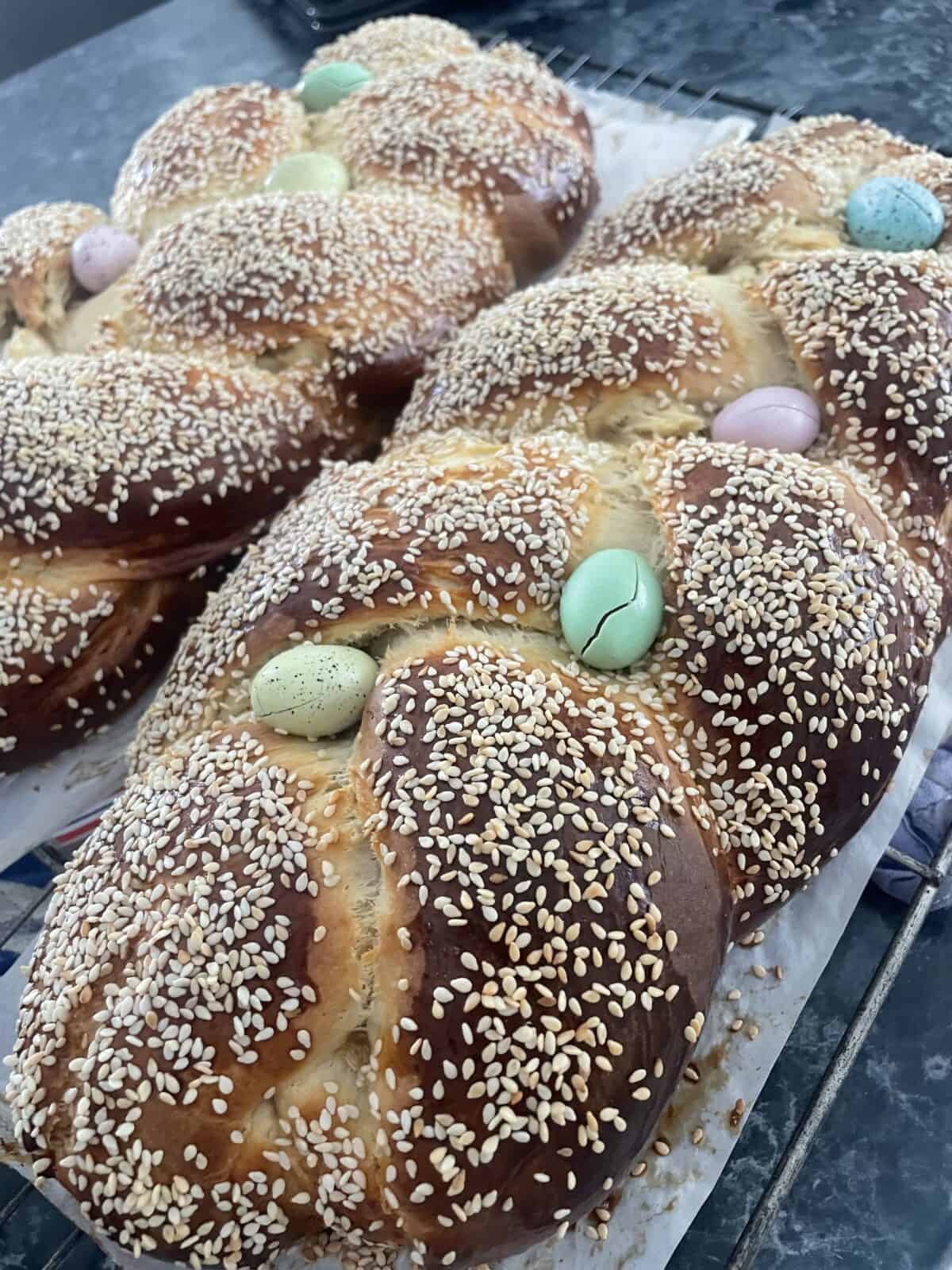 Three braided breads covered with sesame seeds are cooling on a rack. Each loaf has pastel-colored candy eggs nestled in the braids, giving a festive appearance.