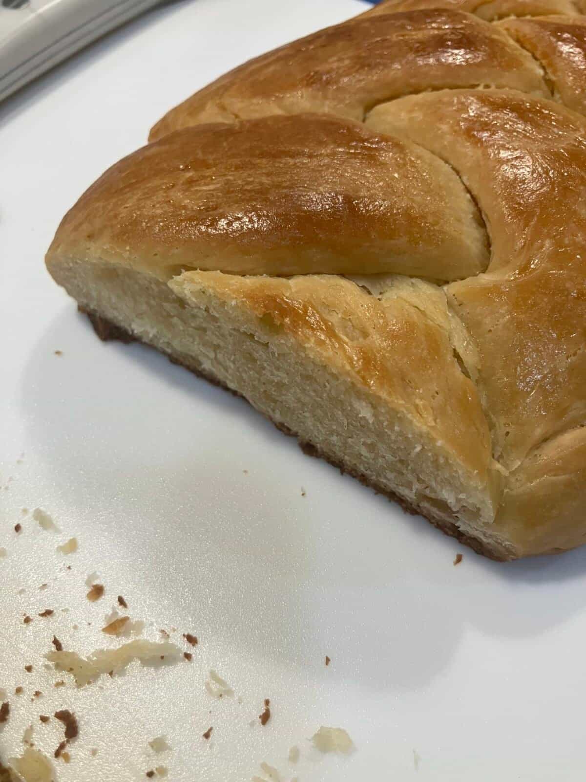 A close-up of a sliced braided loaf of bread on a white surface, with a shiny golden crust and some crumbs scattered nearby.