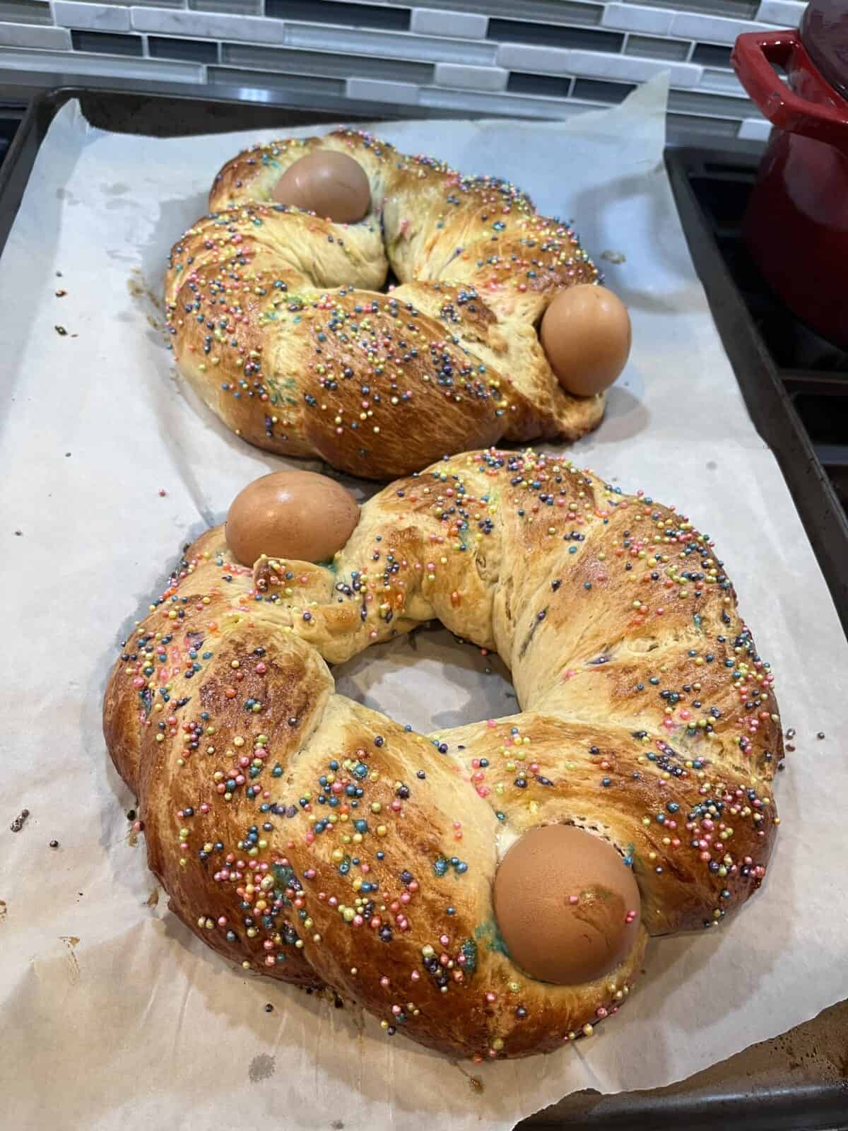 Two braided ring-shaped Sweet Easter Breads topped with colorful sprinkles and whole eggs are baked on parchment paper on a tray, sitting on a kitchen counter with a tiled backsplash in the background.