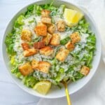 A bowl of Lemon Parmesan Salad topped with croutons, grated parmesan, and lemon wedges, with a gold fork resting inside. The bowl sits on a white surface with a white cloth nearby.