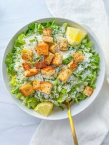 A bowl of Lemon Parmesan Salad topped with croutons, grated parmesan, and lemon wedges, with a gold fork resting inside. The bowl sits on a white surface with a white cloth nearby.