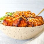 A speckled ceramic bowl filled with grilled chicken pieces and orange sauce, served over rice with green vegetables, evokes the comfort of a classic Salmon Rice Bowl. A chopstick rests inside the bowl on a light background.