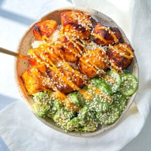 A Salmon Rice Bowl featuring white rice topped with glazed, grilled salmon cubes, sliced cucumber salad, sesame seeds, and a drizzle of orange sauce, placed on a white cloth napkin with sunlight streaming in.