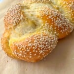 A close-up of a golden-brown braided Sweet Easter Bread, topped with sesame seeds and resting on a sheet of parchment paper.