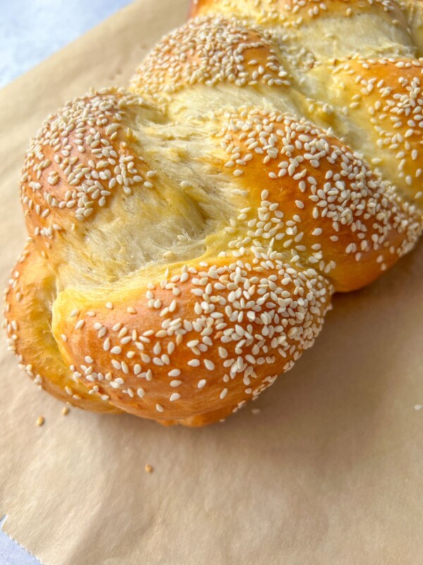 A close-up of a golden-brown braided Sweet Easter Bread, topped with sesame seeds and resting on a sheet of parchment paper.