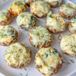 A plate of stuffed mushrooms topped with melted cheese and herbs, arranged neatly on a white dish. The filling appears creamy with bits of green leafy vegetables.