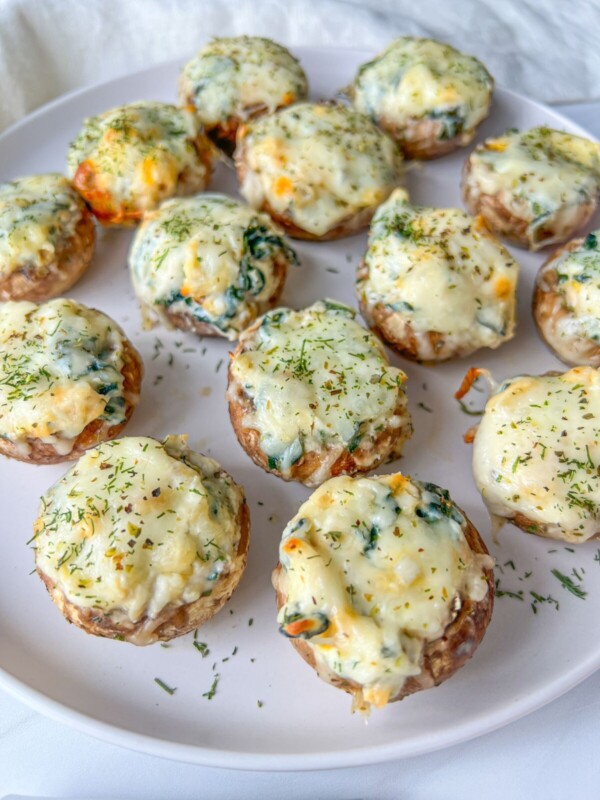 A plate of stuffed mushrooms topped with melted cheese and herbs, arranged neatly on a white dish. The filling appears creamy with bits of green leafy vegetables.