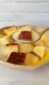 A spoon holds a piece of sweet bread with milk-soaked toast, while more bread pieces and creamy milk rest in a speckled bowl beneath on a white surface.