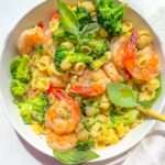 A bowl of Shrimp and Broccoli Pasta features tender pasta shells, broccoli florets, and shrimp, all garnished with fresh basil leaves. Served on a white surface with a gold fork and a white napkin nearby.