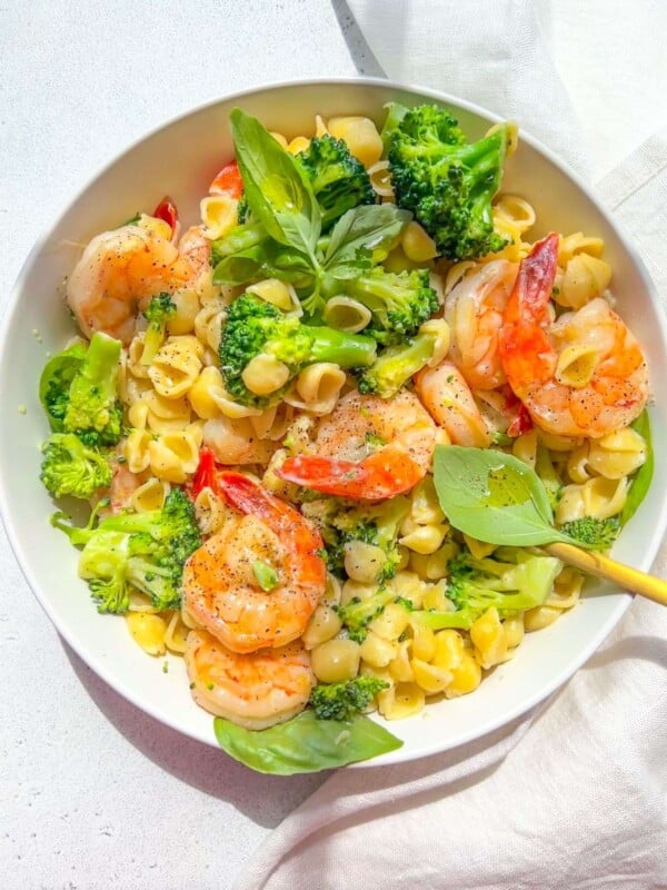A bowl of Shrimp and Broccoli Pasta features tender pasta shells, broccoli florets, and shrimp, all garnished with fresh basil leaves. Served on a white surface with a gold fork and a white napkin nearby.