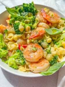 A bowl of Shrimp and Broccoli Pasta featuring pasta shells, fresh basil leaves, and a sprinkle of black pepper, served in a white dish on a bright surface.