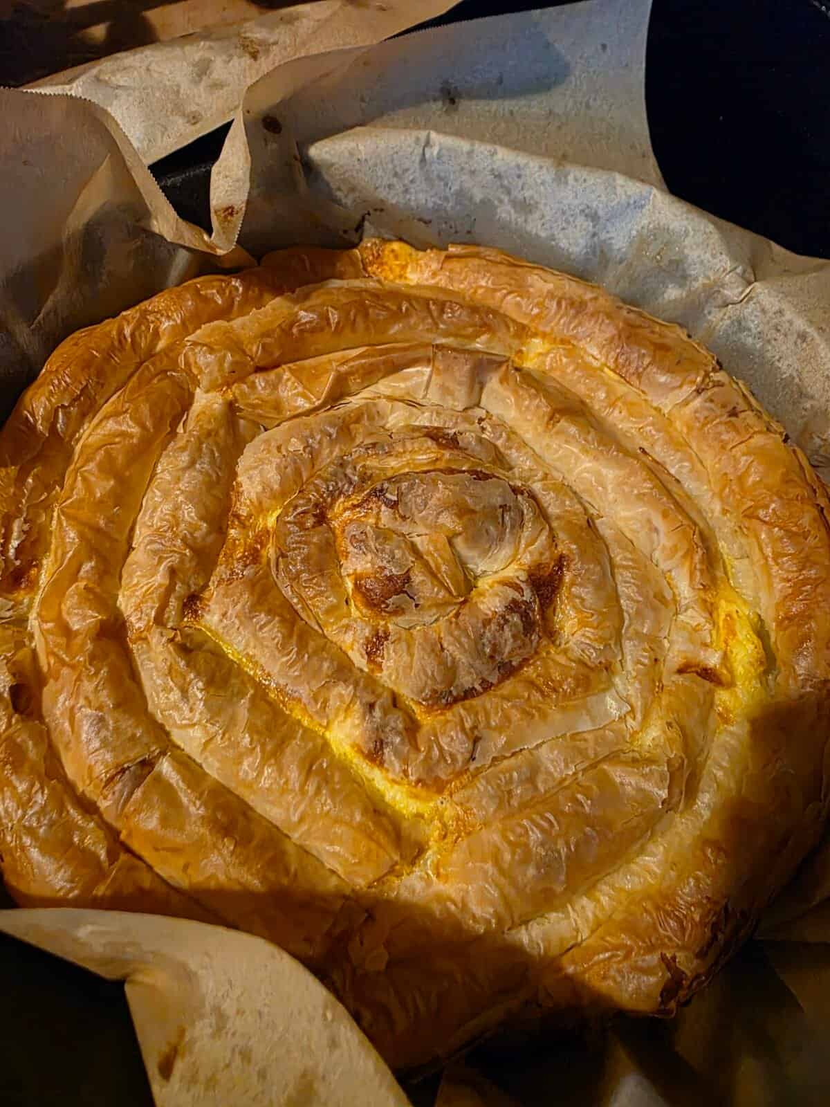 A freshly baked spiral-shaped pie with golden, flaky pastry layers, resting on parchment paper in a baking tray. The surface is crisp and browned, indicating it has just come out of the oven.