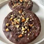 Two round Chocolate Covered Rice Cakes topped with chopped nuts and chocolate pieces sit on a white plate with a light background.