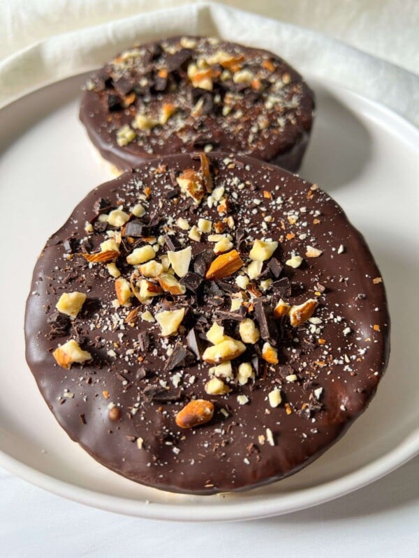 Two round Chocolate Covered Rice Cakes topped with chopped nuts and chocolate pieces sit on a white plate with a light background.