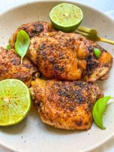 A plate with three pieces of seasoned, grilled lime chicken thighs, garnished with fresh basil leaves and halved limes sprinkled with chili powder. This vibrant Lime Chicken Recipe is perfectly presented with a gold fork resting beside the food.