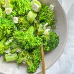 A bowl of fresh sauteed broccoli with feta, mixed with sliced garlic and small cubes of white cheese, served on a light-colored plate with a gold fork resting inside.