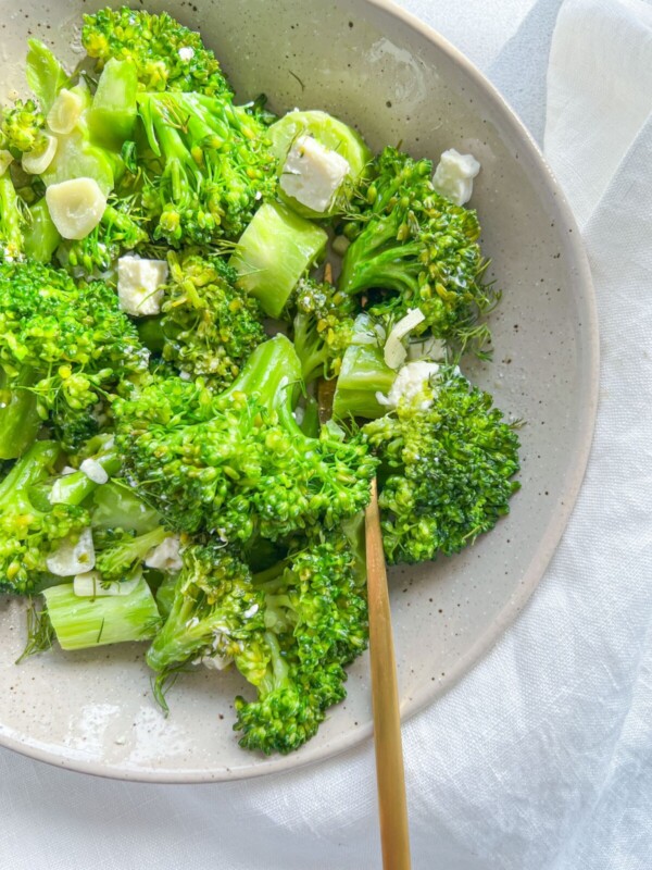 A bowl of fresh sauteed broccoli with feta, mixed with sliced garlic and small cubes of white cheese, served on a light-colored plate with a gold fork resting inside.