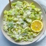 A bowl of chopped romaine lettuce forms the base of this classic Caesar Salad, topped with grated cheese and cracked pepper, garnished with a halved lemon, set on a light surface with a wooden fork and white napkin nearby.