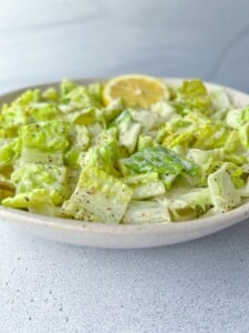 A close-up of a bowl of chopped romaine lettuce tossed in a creamy Green Goddess dressing, garnished with black pepper and a slice of lemon on the rim. The background is a light gray textured surface.