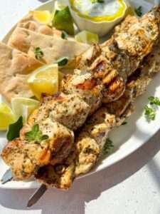 Chicken Souvlaki Skewers with herbs are served on a plate with pita bread, lemon wedges, a small bowl of creamy dip, and fresh herbs, all beautifully arranged in bright natural light.