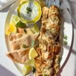 Chicken Souvlaki Skewers are served on a white plate with pita bread, lemon and lime wedges, and a bowl of tzatziki sauce, all garnished with fresh herbs in bright natural light.