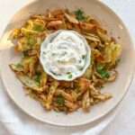 A beige plate with crispy vegetable chips garnished with herbs surrounds a bowl of creamy white dip, set on a white surface with a white napkin underneath—perfect alongside Greek Lemon Potatoes for a flavorful spread.