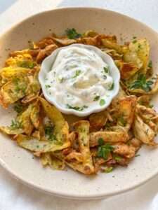 A plate of crispy potato skins inspired by Greek Lemon Potatoes is garnished with fresh herbs, surrounding a small bowl of creamy white dipping sauce, also topped with chopped herbs.
