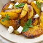 A plate of golden, seasoned Greek Lemon Potatoes wedges garnished with fresh herbs and small cubes of white cheese, with a gold fork on the side.
