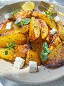 A bowl of golden Greek Lemon Potatoes wedges garnished with fresh herbs, lemon wedges, and small cubes of feta cheese, with a gold fork resting on the side.