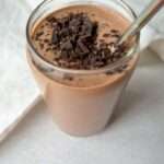 A glass filled with a healthy chocolate smoothie, topped with chopped dark chocolate pieces and a metal straw, sits on a light surface near a white cloth.