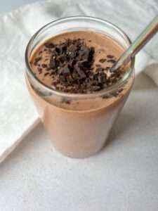 A glass filled with a healthy chocolate smoothie, topped with chopped dark chocolate pieces and a metal straw, sits on a light surface near a white cloth.
