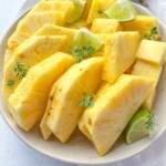 A plate of fresh, sliced pineapple—showcasing how to cut pineapple—arranged with lime wedges and garnished with small green herbs on a light-colored surface.