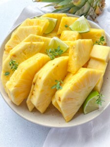 A plate of fresh, sliced pineapple—showcasing how to cut pineapple—arranged with lime wedges and garnished with small green herbs on a light-colored surface.
