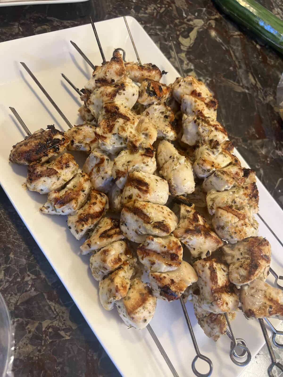 Grilled chicken skewers with spices are arranged on metal sticks and served on a rectangular white plate, placed on a dark marble countertop.