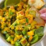 A close-up of a hand holding a chip topped with mango salsa, featuring diced mango, avocado, herbs, and spices. A bowl of the fresh Mango Salsa Recipe and a lime wedge are visible in the background.