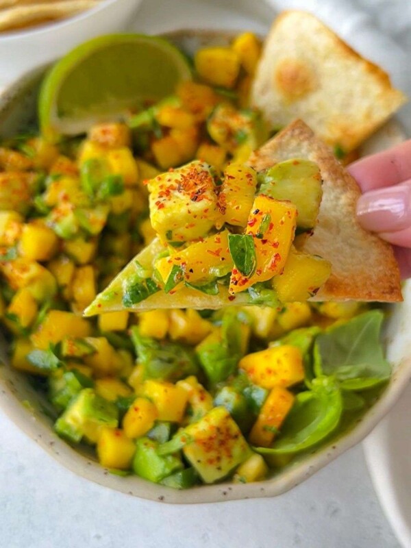 A close-up of a hand holding a chip topped with mango salsa, featuring diced mango, avocado, herbs, and spices. A bowl of the fresh Mango Salsa Recipe and a lime wedge are visible in the background.