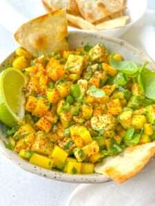 A bowl of vibrant mango avocado salsa, crafted from a delicious Mango Salsa Recipe, garnished with chili powder, fresh herbs, and a lime wedge, served alongside crispy tortilla chips.
